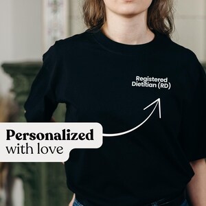 Pu&ograve; includere: T-shirt nero girocollo con la scritta "Registered Dietitian (RD)" e una freccia verso l'alto. La frase "Personalizzato con amore" &egrave; visualizzata in un fumetto bianco.