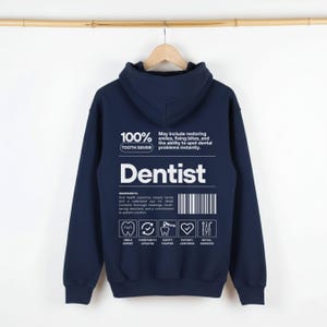 Puede incluir: Sudadera con capucha azul marino con la palabra "Dentist" en blanco. La parte posterior de la sudadera con capucha presenta el texto "100% Tooth Saver" y una lista de "ingredientes" con iconos. La sudadera con capucha está colgada en una percha de madera.