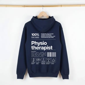 May include: A navy blue hoodie with the words "Physio therapist" printed in white. The back of the hoodie has a graphic with the text "100% Pain Tamer" and a list of ingredients and icons. The hoodie is suspended from a wooden hanger.