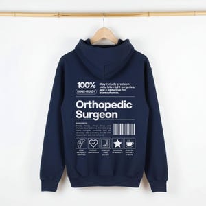 Könnte beinhalten: Ein marineblauer Kapuzenpullover mit weißem Text und Grafiken auf der Rückseite. Das Design enthält die Worte Orthopedic Surgeon, einen Strichcode sowie Symbole für Knochenarchitektur, Patientenorientierung, Mobilität und Kaffee.