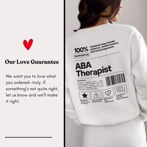 K&ouml;nnte beinhalten: Wei&szlig;es Sweatshirt mit dem Text "ABA Therapist" in Schwarz. Auf der R&uuml;ckseite des Sweatshirts befinden sich ein Barcode und Symbole. Der Text "Our Love Guarantee" wird mit einem roten Herzen angezeigt.