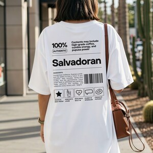 Pu&ograve; includere: T-shirt bianco con la parola "Salvadoran" in grassetto nero. La maglietta presenta il testo "100% Autentico" e un elenco di ingredienti. Una borsa marrone &egrave; appesa alla spalla della persona che indossa la maglietta.