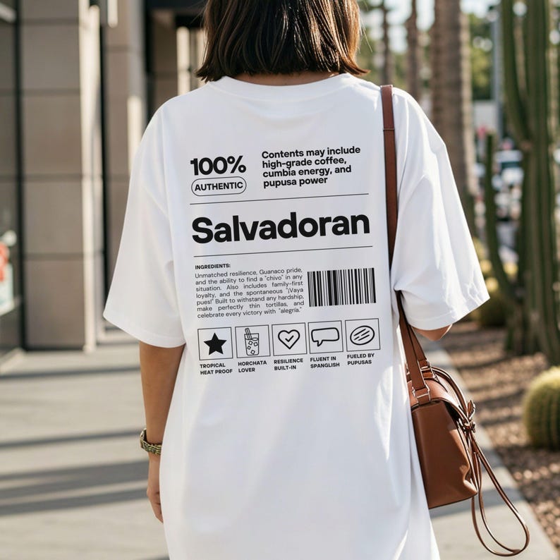 Pu&ograve; includere: T-shirt bianco con la parola "Salvadoran" in grassetto nero. La maglietta presenta il testo "100% Autentico" e un elenco di ingredienti. Una borsa marrone &egrave; appesa alla spalla della persona che indossa la maglietta.