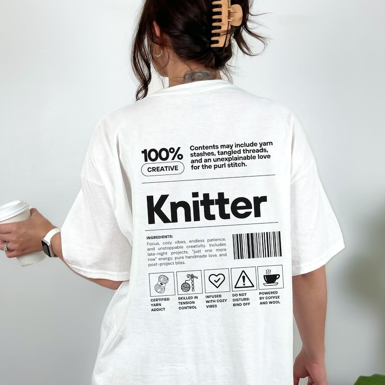 Op de afbeelding: Wit T-shirt met het woord "Knitter" in grote zwarte letters. Het shirt heeft de tekst "100% Creatief" en "Inhoud kan garenvoorraden, verwarde draden en een onverklaarbare liefde voor de averechtse steek bevatten."