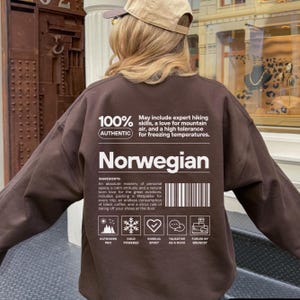 Könnte beinhalten: Ein braunes Sweatshirt mit dem Wort "Norwegian" in Weiß. Das Kleidungsstück enthält Text, der norwegische Eigenschaften und Symbole beschreibt. Der Text lautet "100% Authentic" und weitere Informationen.