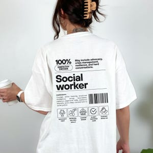 Könnte beinhalten: Weißes T-Shirt mit dem Wort „Social Worker“ in fetter schwarzer Schrift. Das Shirt zeigt eine Grafik mit dem Text „100% Empathy Driven“ und eine Zutatenliste, einschließlich Mut und Empathie. Zusätzliche Symbole sind ebenfalls vorhanden.