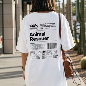 Op de afbeelding: Wit t-shirt met de tekst "Animal Rescuer" in het zwart. Het shirt bevat een lijst met ingredi&euml;nten en pictogrammen met betrekking tot dierenredding. Het shirt wordt gedragen door een persoon met een bruine handtas.