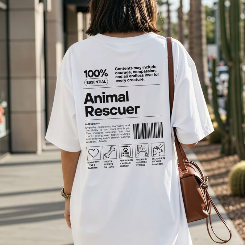 Op de afbeelding: Wit t-shirt met de tekst "Animal Rescuer" in het zwart. Het shirt bevat een lijst met ingredi&euml;nten en pictogrammen met betrekking tot dierenredding. Het shirt wordt gedragen door een persoon met een bruine handtas.
