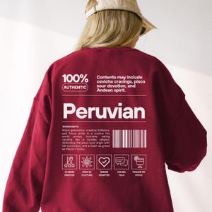 May include: A maroon crewneck sweatshirt featuring white text on the back that reads 100% AUTHENTIC, Peruvian, and lists ingredients including ceviche cravings and pisco sour devotion, alongside a barcode and five icons labeled Cuisine Master, Rich in Culture, Warm Hearted, Causa Talk, and Fueled by Pisco.