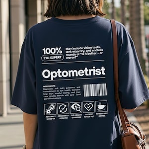 May include: Navy blue t-shirt with white text and graphics. The shirt features the word "Optometrist" and phrases like "Eye-Expert" and "Powered by Coffee." Additional text includes "Accuracy Mode On" and "Patient Focus 24/7."