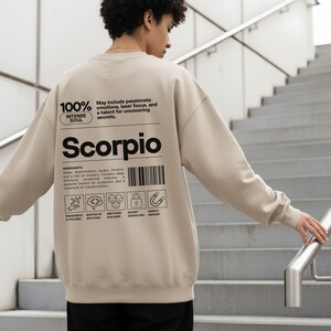 Pode incluir: Camisola bege com a palavra "Scorpio" impressa em preto. A parte de tr&aacute;s da camisola apresenta texto sobre os tra&ccedil;os de Escorpi&atilde;o, incluindo "alma intensa" e "emo&ccedil;&otilde;es apaixonadas". O design inclui um c&oacute;digo de barras e v&aacute;rios &iacute;cones.