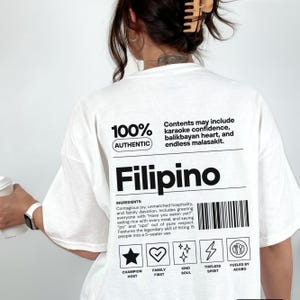 Könnte beinhalten: Weißes T-Shirt mit dem Wort "Filipino" in fetter schwarzer Schrift. Das Shirt enthält Text über die philippinische Kultur, wie Karaoke-Selbstvertrauen und unübertroffene Gastfreundschaft. Das Shirt hat auch den Text "100% Authentic".