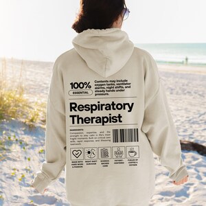 Może przedstawiać: Beżowa bluza z kapturem z czarnym napisem "Respiratory Therapist". Z tyłu bluzy znajduje się kod kreskowy oraz tekst "100% Essential" i "Contents may include oxygen tanks, ventilator alarms, night shifts, and steady hands under pressure."
