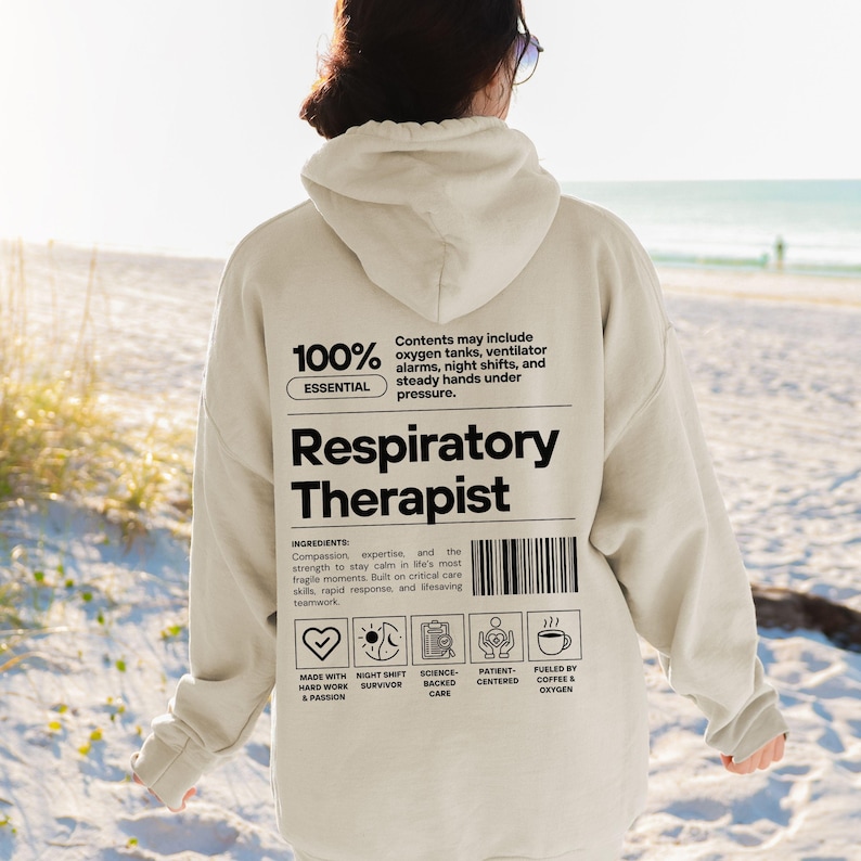Może przedstawiać: Beżowa bluza z kapturem z czarnym napisem "Respiratory Therapist". Z tyłu bluzy znajduje się kod kreskowy oraz tekst "100% Essential" i "Contents may include oxygen tanks, ventilator alarms, night shifts, and steady hands under pressure."