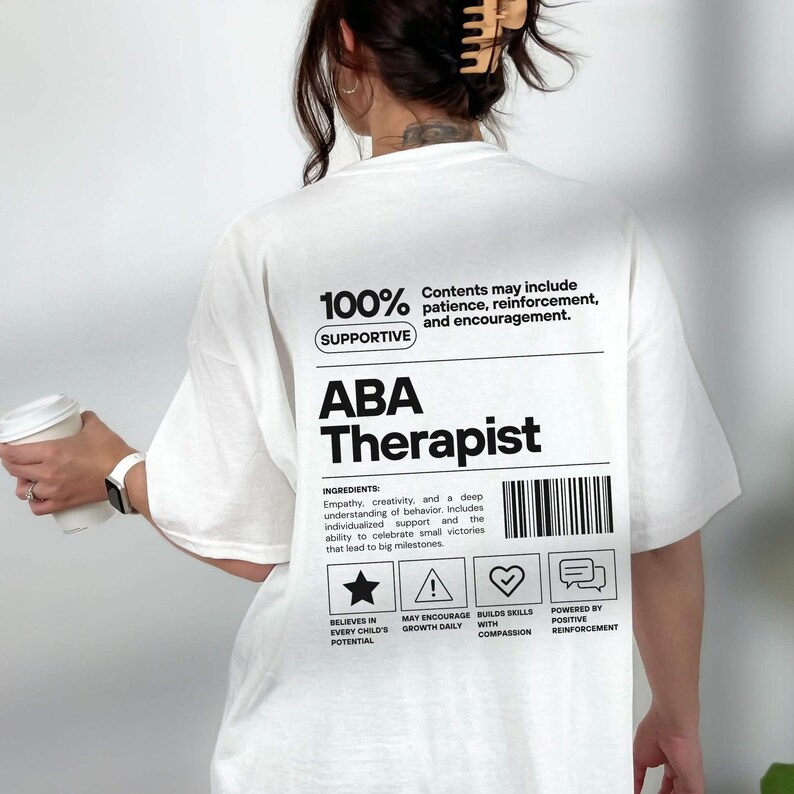 K&ouml;nnte beinhalten: Wei&szlig;es T-Shirt mit dem Text "ABA Therapist" in Schwarz. Das Shirt enth&auml;lt zus&auml;tzlichen Text: "100% Supportive" und "Contents may include patience, reinforcement, and encouragement." Das Shirt hat auch einen Barcode und Symbole.