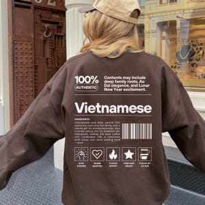 May include: A brown sweatshirt with the word "Vietnamese" in white block letters. The back of the sweatshirt has text that reads "100% Authentic" and a list of ingredients. The sweatshirt also has icons and text that read "Stay Strong", "Warm Hearted", "Flavor Expert", "Heritage Proud", and "Fueled by Cà Phê".