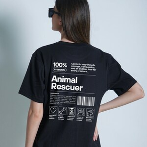 Op de afbeelding: Zwart t-shirt met de witte tekst "Animal Rescuer". Het shirt bevat een lijst met "ingredi&euml;nten" en pictogrammen met betrekking tot dierenredding. De tekst "100% Essential" staat er ook op.