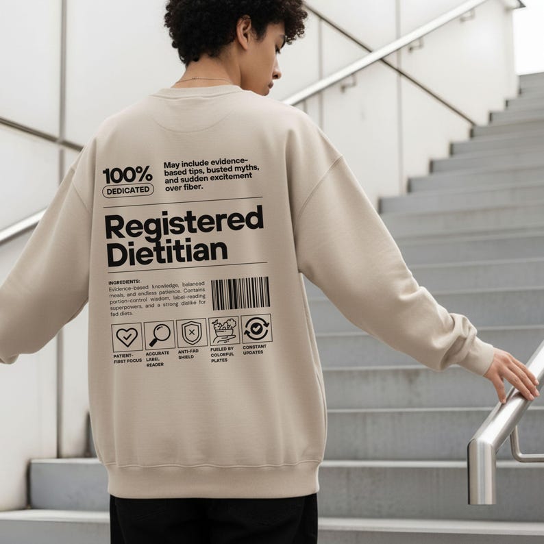 Pu&ograve; includere: Felpa beige con la scritta "Registered Dietitian" in nero. Il retro della felpa presenta un design di etichetta nutrizionale con icone e testo, tra cui "100% Dedicated". La felpa &egrave; di colore neutro.