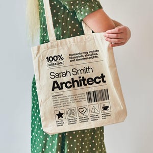 May include: A cream-colored tote bag with the text "100% Creative" and "Sarah Smith Architect" printed in black. The bag includes a barcode and various icons. The text "Contents may include blueprints, sketches, and sleepless nights" is also printed on the bag.