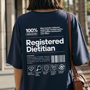Pu&ograve; includere: T-shirt blu navy con testo bianco che recita "Registered Dietitian". La maglietta include un elenco di "ingredienti" e icone. Il testo "100% Dedicated" &egrave; in alto. &Egrave; visibile una borsa a tracolla in pelle marrone.