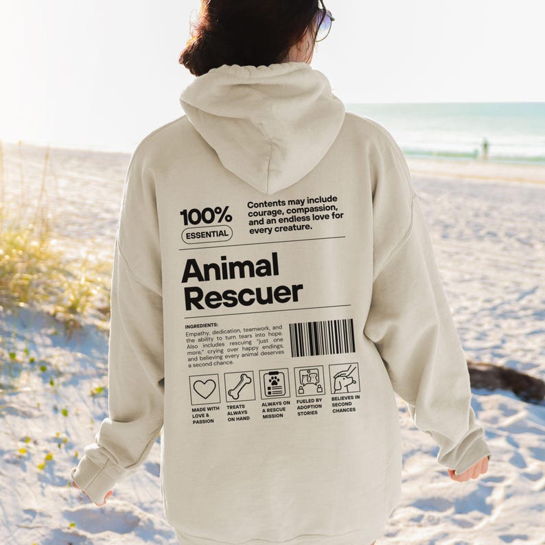 Op de afbeelding: Beige hoodie met de tekst "Animal Rescuer" in het zwart. De achterkant van de hoodie heeft een lijst met ingredi&euml;nten en pictogrammen die betrekking hebben op dierenredding. De hoodie is gemaakt van zacht materiaal.