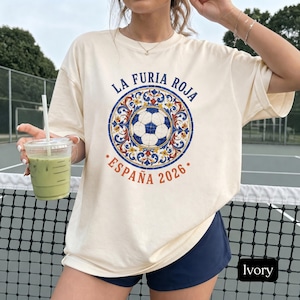 May include: An ivory short-sleeve t-shirt featuring a central circular graphic with a soccer ball surrounded by blue, red, and yellow floral patterns. The text reads LA FURIA ROJA and ESPAÑA 2026 in blue and orange lettering around the design.