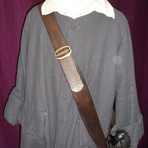 Peut inclure: Une ceinture de sabre en cuir marron avec une boucle en laiton portée sur un manteau gris. La ceinture est portée en diagonale sur la poitrine et a une épée attachée à l'extrémité.