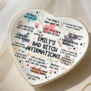 May include: Heart-shaped ceramic trinket dish with gold trim, featuring the text "Emily's Bad Bitch Affirmations" and various empowering phrases. The dish is white with colorful accents and floral designs.