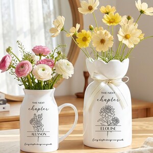May include: Two white ceramic vases with floral arrangements. One vase has a handle and contains pink and white flowers. The other vase has a scalloped top and contains yellow flowers. Both vases have the text "THE NEXT chapter" and a name below.