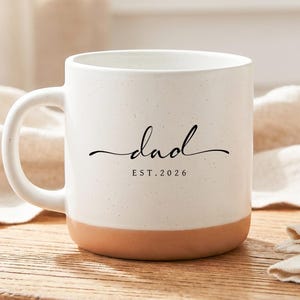 May include: White ceramic mug with a speckled finish and a tan base. The word "dad" is written in a cursive font, with "EST. 2026" below it. The mug is on a wooden surface.