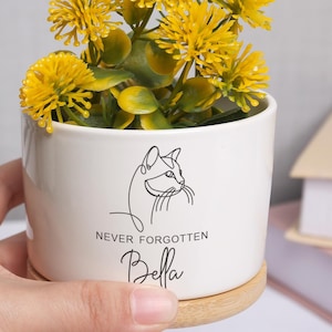 May include: A white ceramic planter with a cat outline and the text "NEVER FORGOTTEN Bella" holds yellow flowers. The planter sits on a small wooden coaster. A hand gently holds the planter.