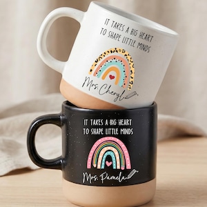 May include: Two ceramic mugs stacked vertically. The top mug is white with a speckled finish, featuring a rainbow graphic, a pencil icon, and the text "It takes a big heart to shape little minds" and "Mrs. Cheryl". The bottom mug is black with a similar design and the name "Mrs. Pamela". Both have a tan base.