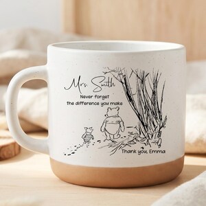 May include: A white ceramic mug with a speckled finish and a tan base. The mug features a black line drawing of Winnie the Pooh and Piglet walking near trees, with the text: Mrs. Smith, Never forget the difference you make, Thank you, Emma.