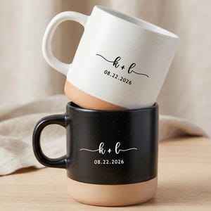 Pode incluir: Duas canecas de cerâmica empilhadas verticalmente, uma branca e uma preta, ambas com textura salpicada e base de argila natural. Cada caneca está impressa com o texto k + b e a data 08.22.2026 num estilo de letra simples.
