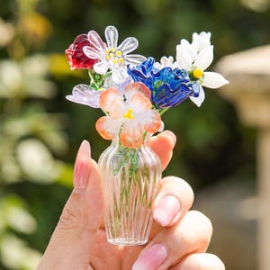 May include: A small glass vase containing a bouquet of colourful glass flowers. The flowers are red, white, blue, and orange. The vase is held in a hand, highlighting the intricate details of the glasswork.