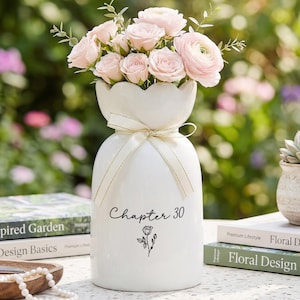 May include: A white ceramic vase with a scalloped rim and a cream-colored ribbon tied around the neck. The vase features the black text "Chapter 30" above a small black rose illustration. It is filled with a bouquet of light pink flowers.