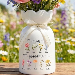 May include: A white ceramic vase with a scalloped top, filled with colorful flowers. The vase has a cream-colored ribbon tied around the neck and features the words "First Mom Now Grandma" with floral illustrations and names.