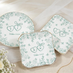 May include: Three white ceramic plates with a floral design and gold trim. Each plate features a pair of connected heart-shaped pendants with initials and a date. The plates are round, square, and a unique shape.