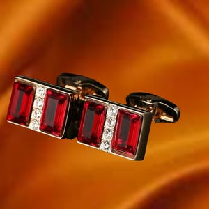 Puede incluir: Un par de gemelos dorados con piedras preciosas rojas rectangulares y pequeñas piedras claras. Los gemelos tienen un diseño clásico, perfectos para ocasiones formales o para añadir un toque de elegancia a cualquier atuendo.