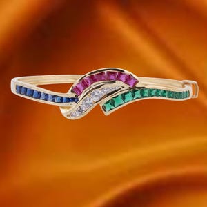 May include: A gold bracelet with a wave design, featuring blue, red, green, and white gemstones. The bracelet has a hinged clasp. The gemstones are rectangular and square cut. The background is orange.