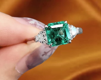 Può includere: Un anello d'argento con una grande pietra di smeraldo centrale a taglio quadrato, affiancata da due diamanti più piccoli a taglio triangolare. Lo smeraldo è di un verde vibrante e i diamanti brillano. L'anello è montato su una fascia delicata.