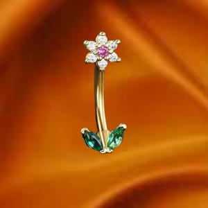 May include: A gold-colored belly button ring with a floral design. The top features a flower with white and pink gemstones. The bottom has two green, teardrop-shaped gems. The curved bar is gold.