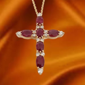 May include: A gold-colored pendant necklace featuring a cross design. The cross is adorned with oval-shaped, deep red gemstones and small, round, clear gemstones. The necklace hangs on a delicate gold chain, set against an orange background.