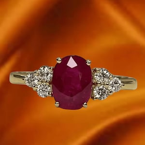 May include: A gold ring featuring a large, oval-cut ruby gemstone as the centerpiece. The ruby is a deep red color. Smaller, round diamonds are clustered on either side of the ruby, adding sparkle. The ring is set against an orange background.