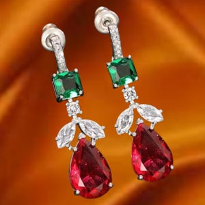 May include: A pair of dangling earrings with a silver-tone metal setting. Each earring features a green square-cut gemstone at the top, followed by a cluster of white, teardrop-shaped stones, and a large, red teardrop-shaped gemstone at the bottom.