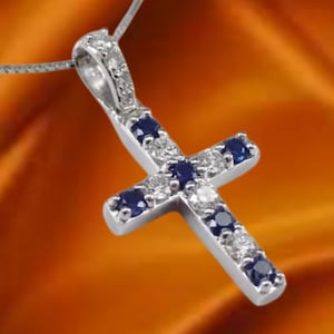 May include: A silver cross pendant with a delicate chain. The cross is adorned with small, round, white and dark blue gemstones. The pendant is set against a warm, orange background, highlighting the jewelry's sparkle and design.