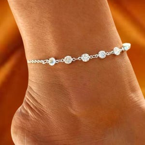 May include: A silver anklet with clear, round, and sparkling gemstones. The anklet is delicate and features a series of gemstones connected by small links. The gemstones are evenly spaced along the chain, creating a simple yet elegant design.