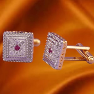 Peut inclure: Une paire de boutons de manchette carrés avec un design argenté et doré. Chaque bouton de manchette présente une pierre précieuse rouge centrale entourée de petites pierres claires. Les bords sont ornés d'une bordure à motifs, et la barre de connexion est dorée.