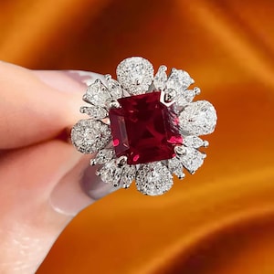 May include: A silver ring with a large, square-cut, deep red gemstone at the centre, encircled by smaller, sparkling white stones. The ring has a floral design. The background is a blurred, warm orange.