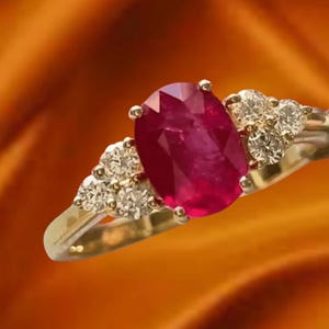 Op de afbeelding: Een gouden ring met een grote, ovaal geslepen robijn als middelpunt. De ring is versierd met kleinere, ronde diamanten aan weerszijden van de robijn, tegen een warme, oranje achtergrond.